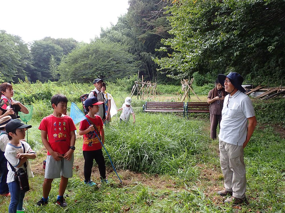 話をする里山をよみがえらせる会会長の荒井さんと話を聞く参加者。背景にはぜかけの木が見える。