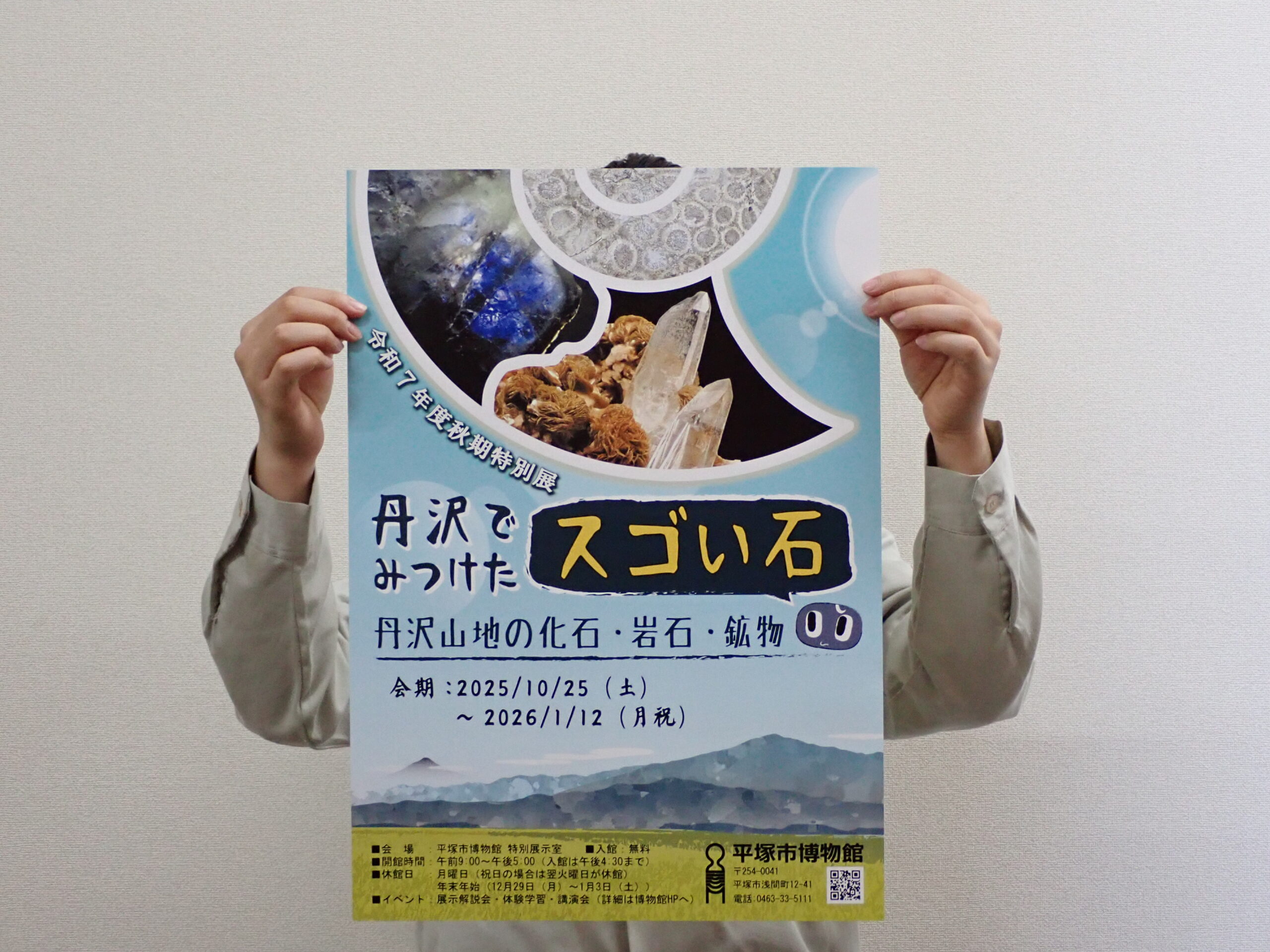 ポスターを正面から撮影したもの。人が持っていて、上半身を覆うくらいの大きさ。B2サイズ。
上部にアンモナイトをイメージした枠があり、特別展で展示される鉱物や化石の画像がはめ込まれている。中央に展示のタイトル、下部に丹沢の山並みのイラストがデザインされている。
