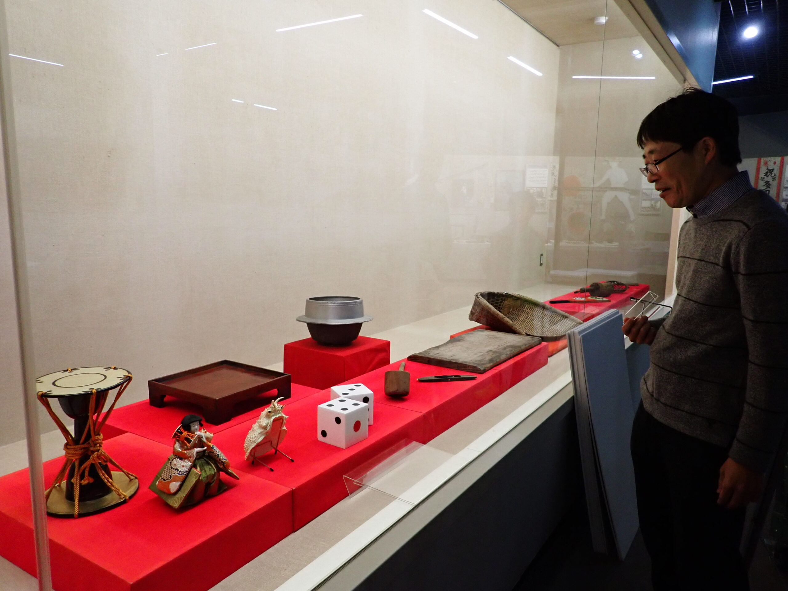 展示ケースの外からケース内の資料を見ているところ。カニの甲羅の他、鼓、サイコロ、羽釜、ひしゃく、機織り機の杼(シャトル)、魅スの外からケース内の資料を見ているところ。カニの甲羅の他、鼓、サイコロ、羽釜、ひしゃく、機織り機の杼(シャトル)、箕、羽子板などが並ぶ。