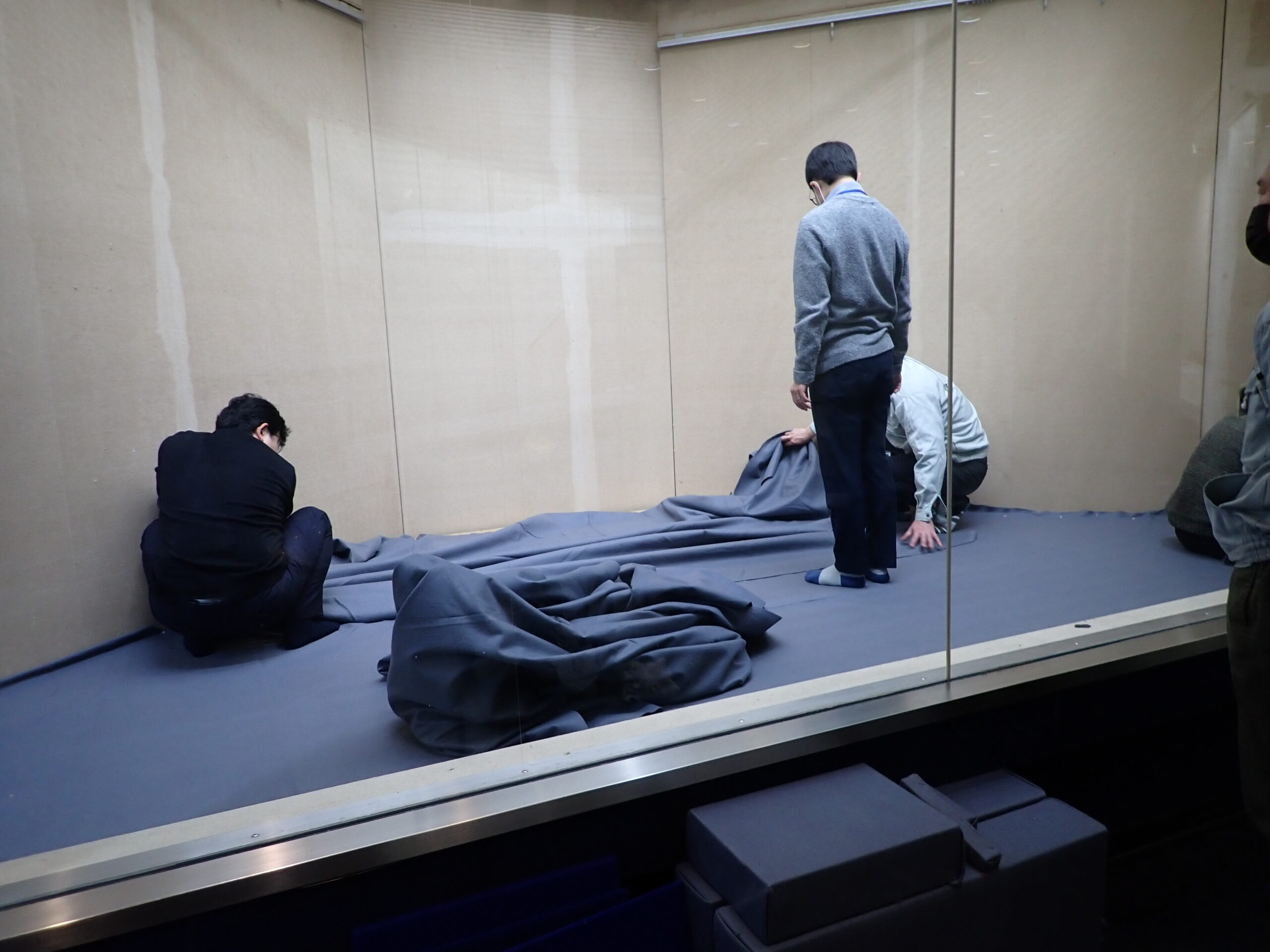 展示ケース内で床面にフェルトを敷く作業をしている様子。幅２ｍ、長さ10ｍ程度のフェルトを切り、前後に並べて敷いている。