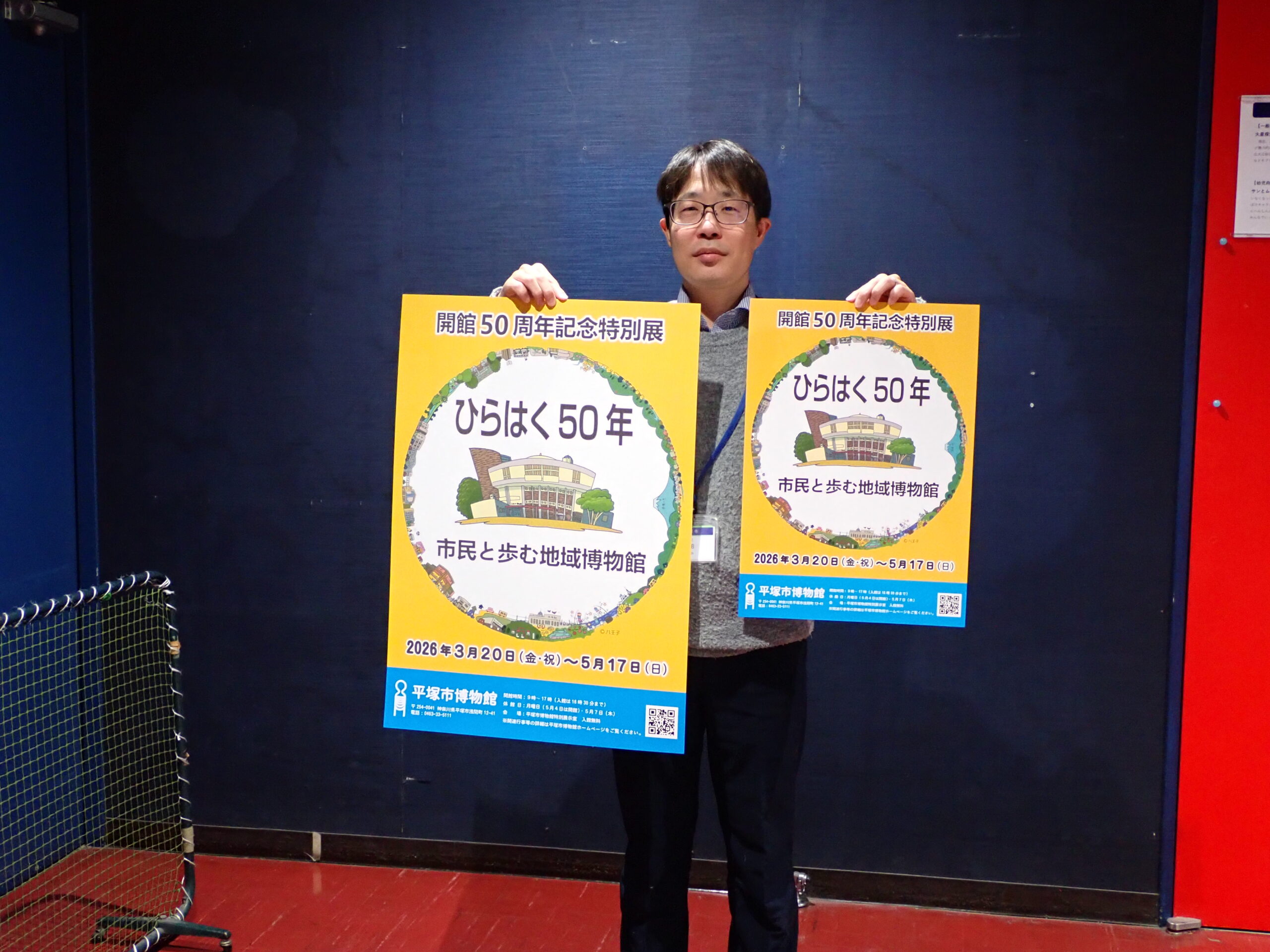 2種類のポスターを持って立つ男性。ポスターの中央に博物館とその周辺の景色がイラストでデザインされている。